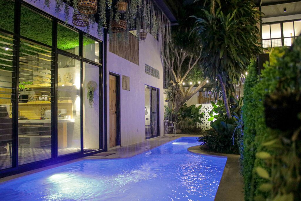 Nighttime view of a narrow backyard pool surrounded by lush greenery and outdoor lighting — time to resurface a pool for both beauty and safety.