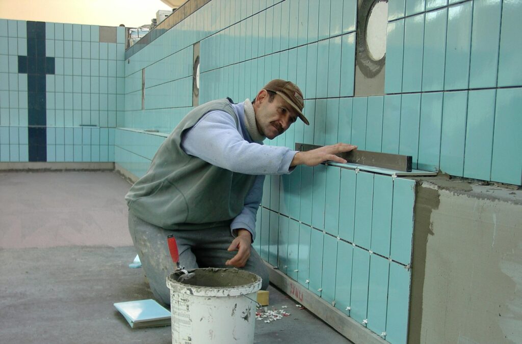 A professional pool builder carefully installing blue ceramic tiles on the interior wall of an empty swimming pool using a level tool.
